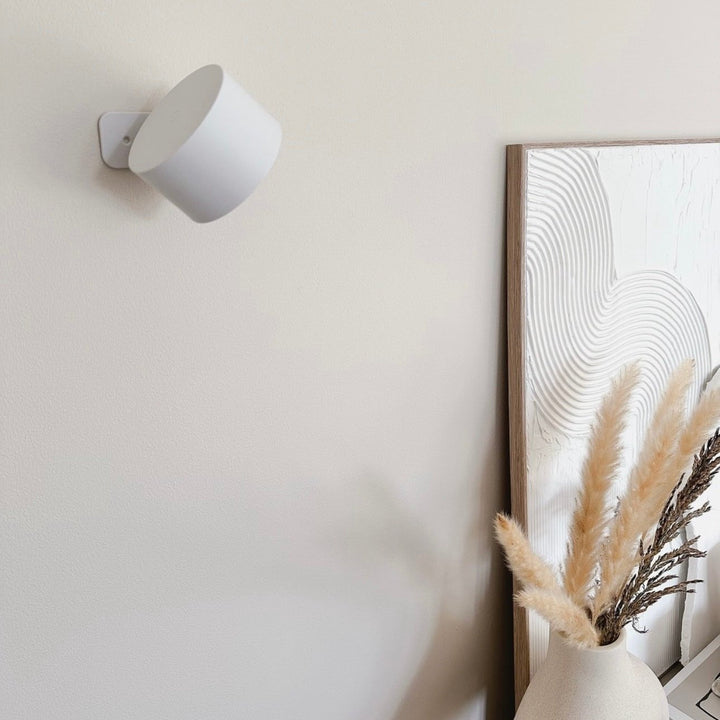White wall light switched off and mounted on a beige wall, next to a textured framed artwork and a vase of pampas grass.