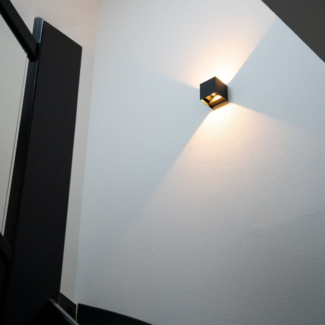 Black cube wall lamp seen from below on a white textured stairwell wall, throwing a warm fan of light upward. 2
