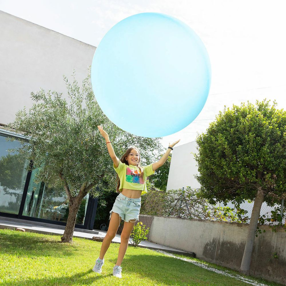 Smiling girl on a backyard lawn tosses a huge translucent blue balloon ball high above her head with both arms raised. 3