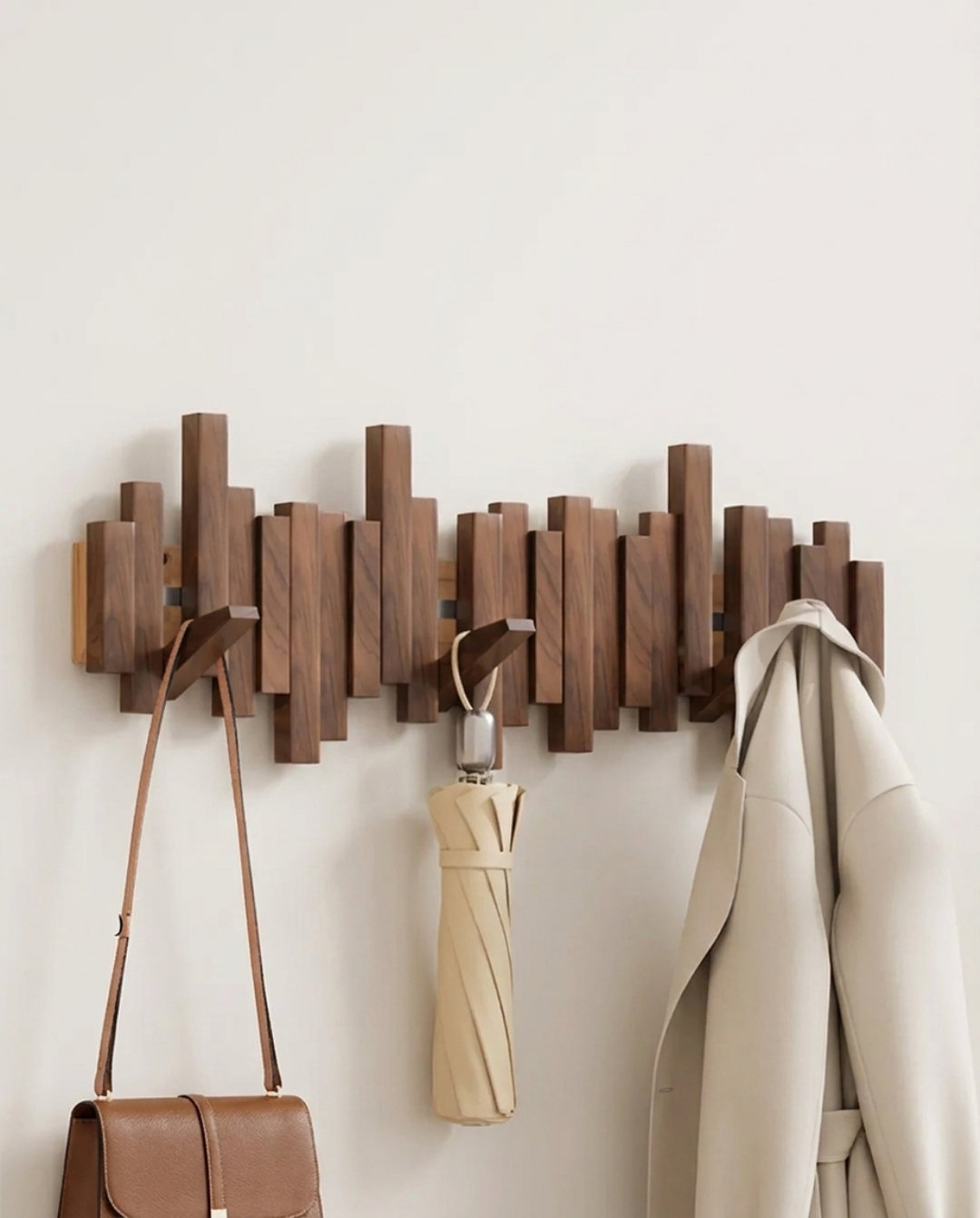 Dark brown wooden wall rack with folding hooks holding a brown leather purse, a beige umbrella and a cream coat.