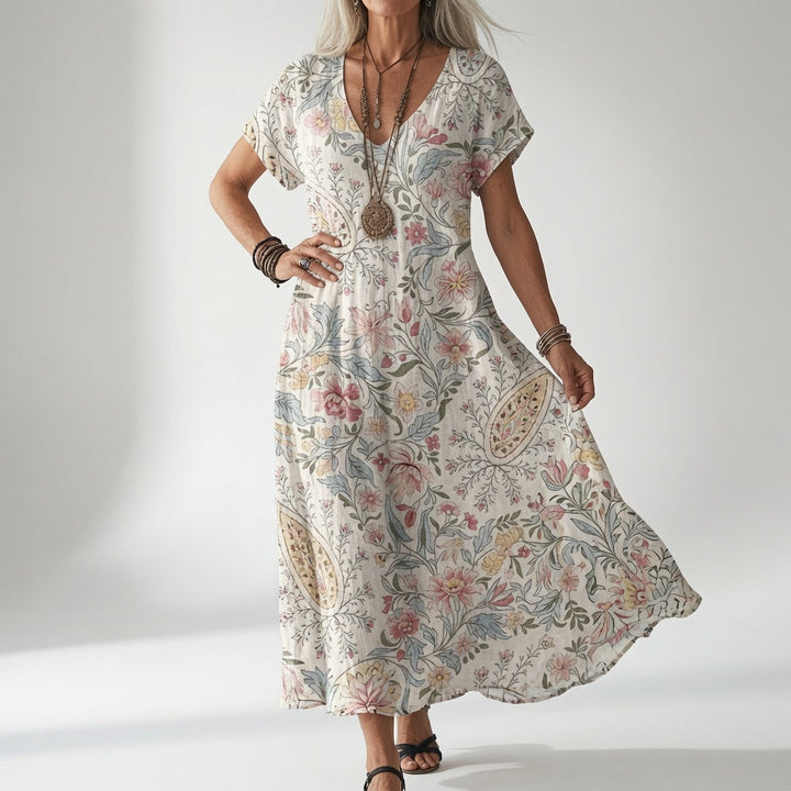 Woman wearing a cream V-neck dress with a pink, blue and yellow paisley floral print, styled with a long beaded necklace and sandals.