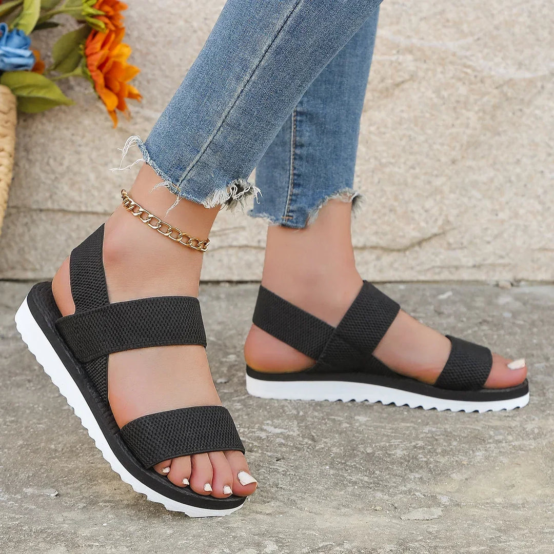 Black elastic double-strap sandals on feet, one angled to show the side, beside orange flowers on a stone floor 3