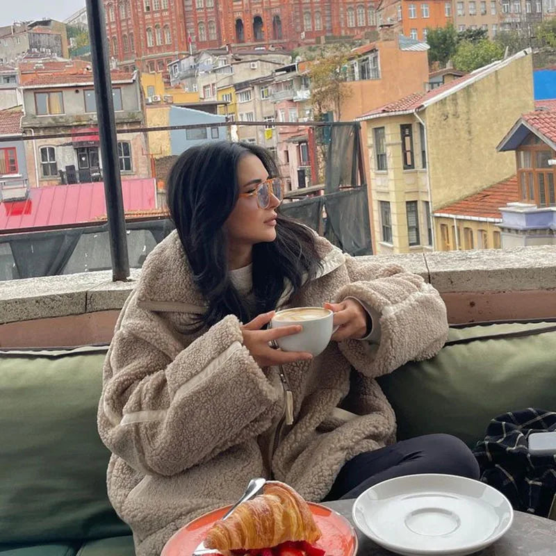Woman in a taupe fleece coat holding a cup of coffee at a rooftop table overlooking buildings 3