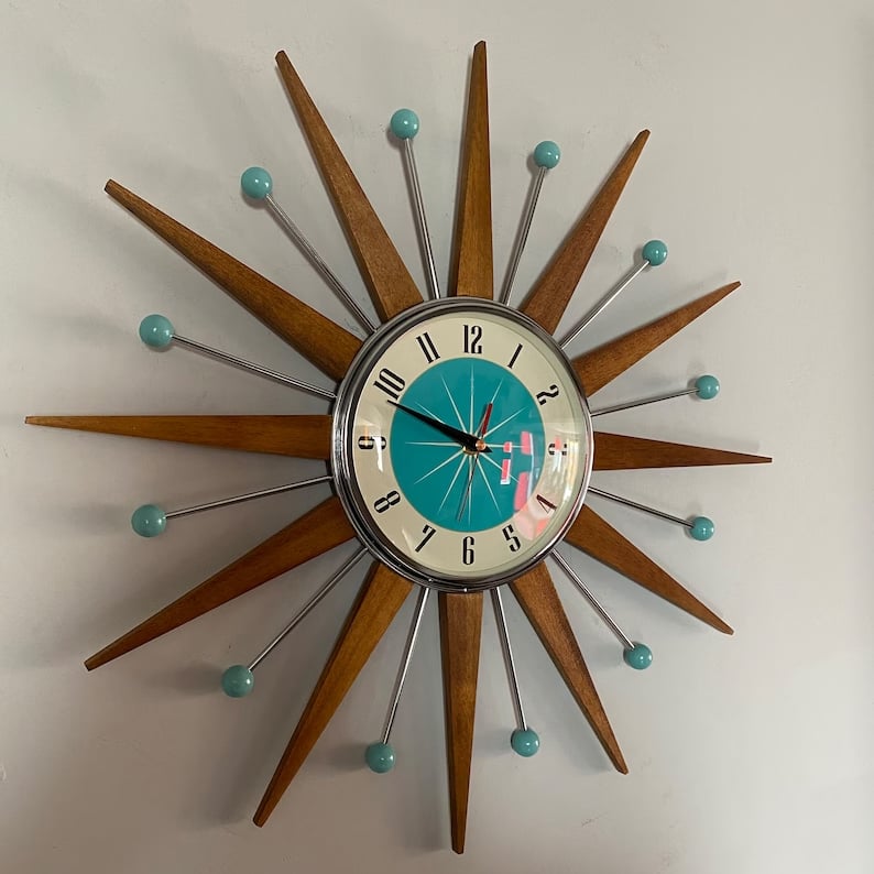 Starburst wall clock detail hanging on a pale gray wall, showing wooden rays, teal ball tips and a teal dial.