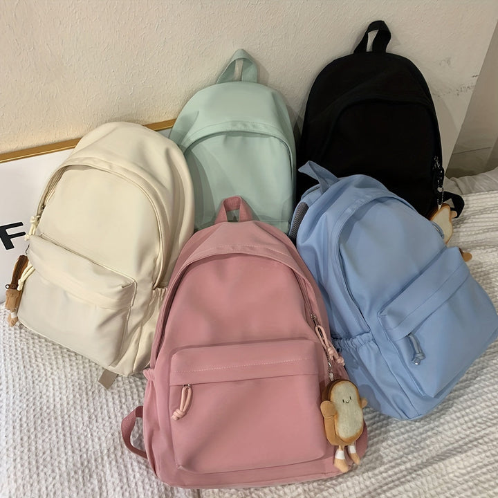 Five backpacks in cream, mint, black, pink and blue resting on a bed, two with small toast-shaped charms attached.
