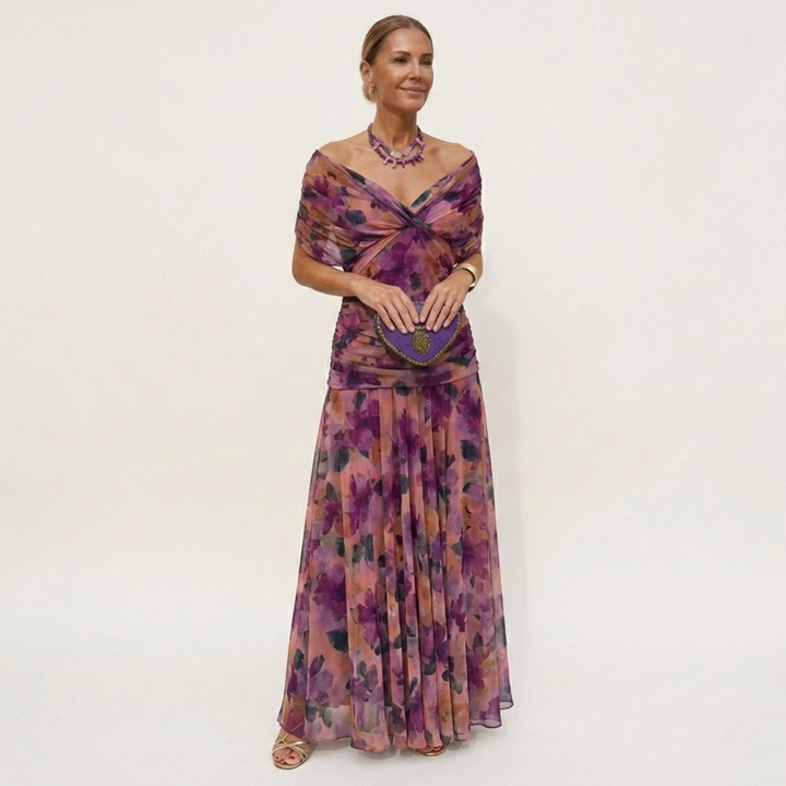 Model in a long off-the-shoulder dress with a purple and peach floral print, twisted bodice and a purple heart-shaped clutch. 4