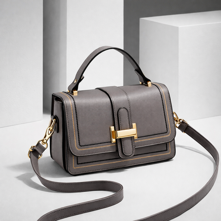 Gray version of the flap bag with gold clasp and contrast stitching, shown at an angle among white blocks. 3