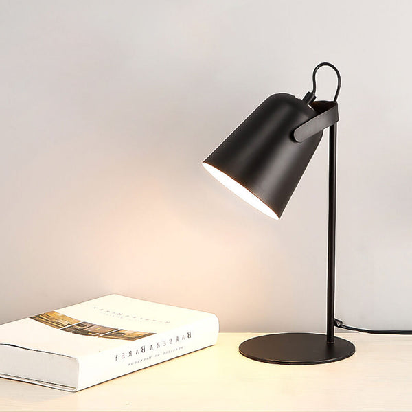 Black version of the lamp lit on a light desk beside a closed book, shade angled left 12