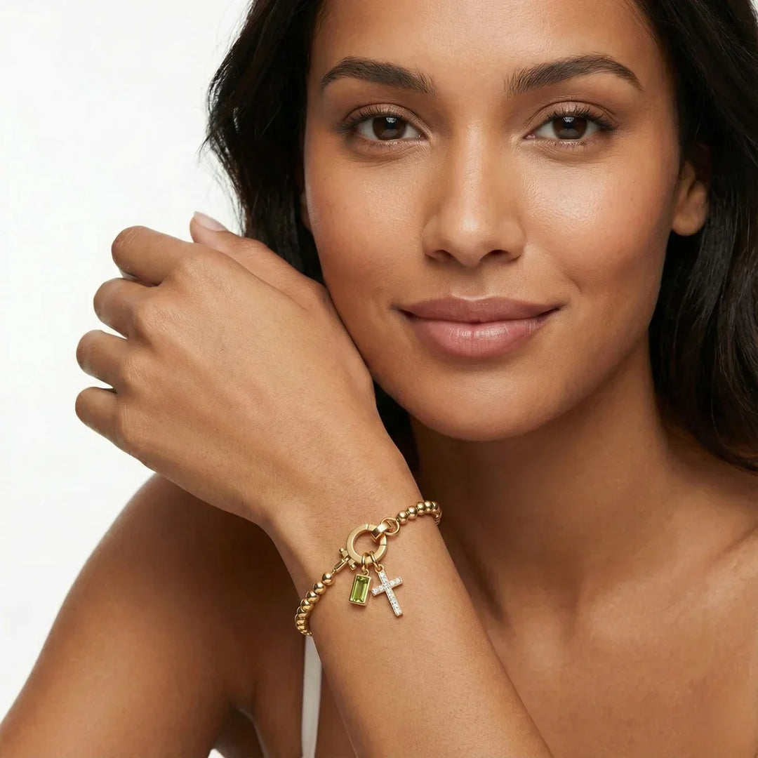 Model smiling with a gold-tone bead bracelet and a green stone charm and crystal cross charm on her wrist.