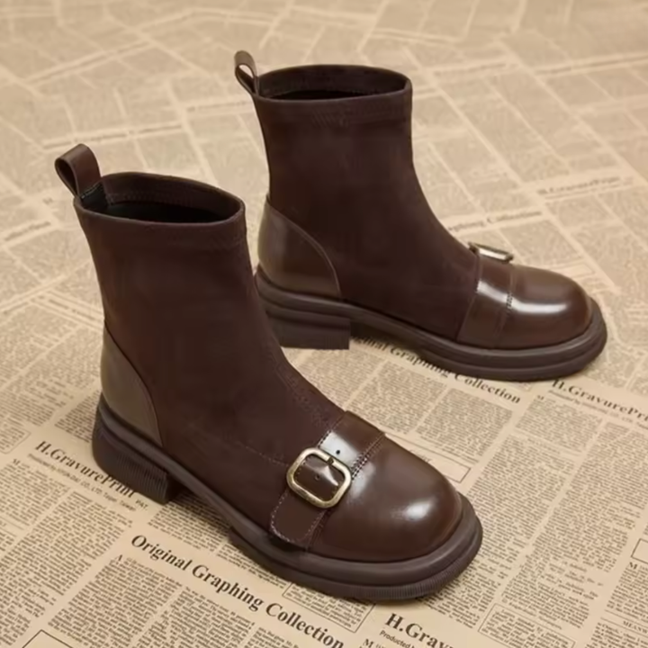 Pair of brown suede boots on a newspaper-print background, showing the buckled leather toe strap, pull tabs and chunky low heel.