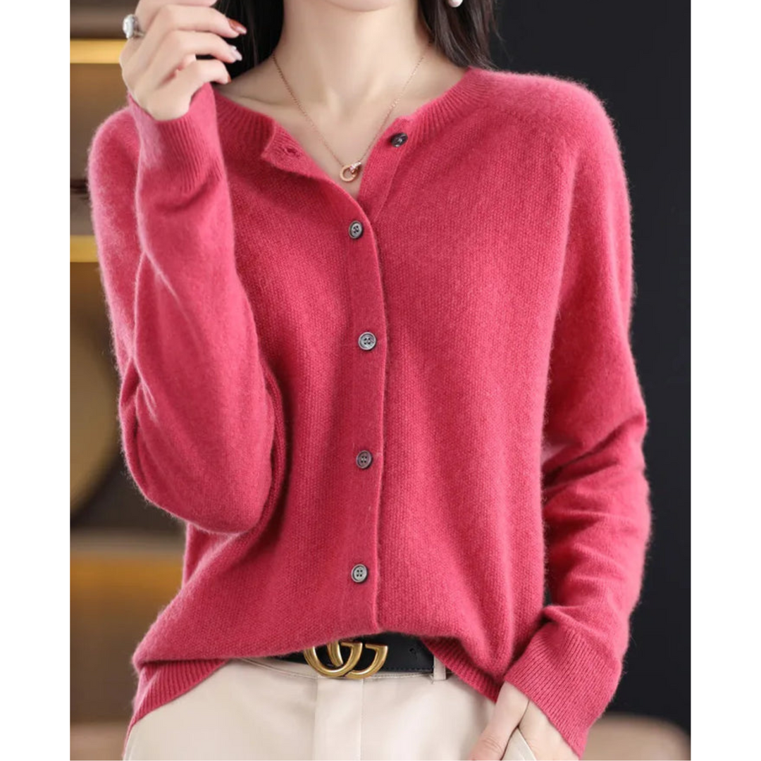 Woman wearing a raspberry pink button-front knit cardigan with round neck, hand raised to her collar, paired with cream trousers and a black belt. 1