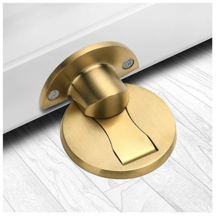 Brushed gold-toned version of the door stopper, shown with its door plate and round floor magnet on light wood flooring. 3