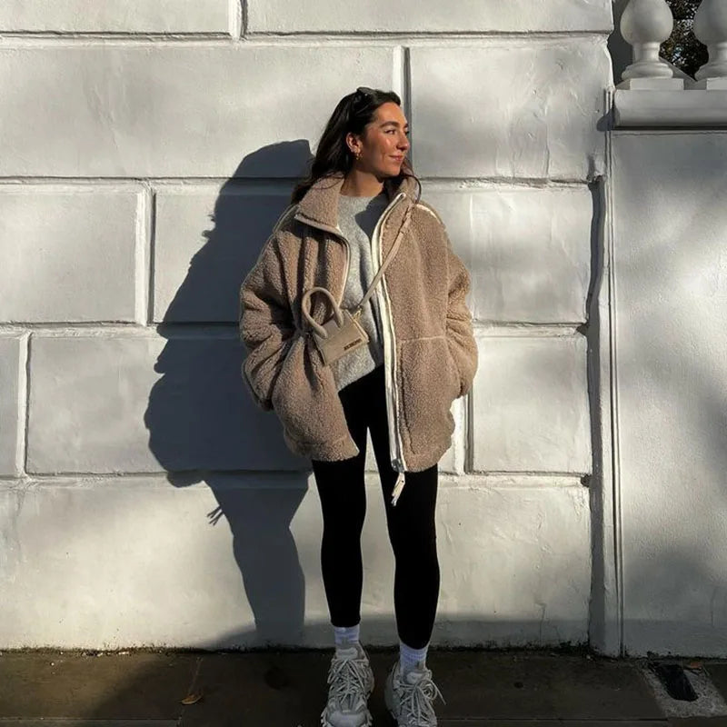Woman in a taupe fleece coat with hands in pockets, black leggings and sneakers, standing by a white wall 4