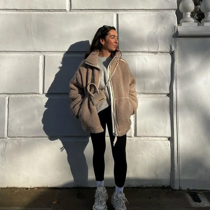 Woman in a taupe fleece coat with hands in pockets, black leggings and sneakers, standing by a white wall 4