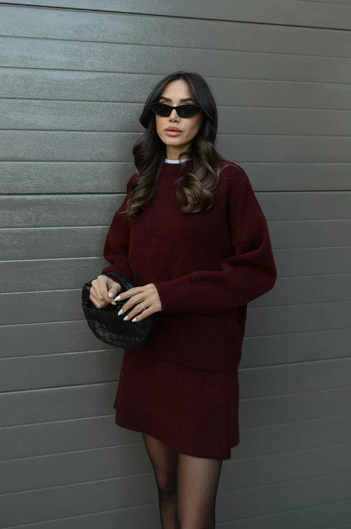 Model leaning against a gray wall in the burgundy knit set, holding a black woven bag 3