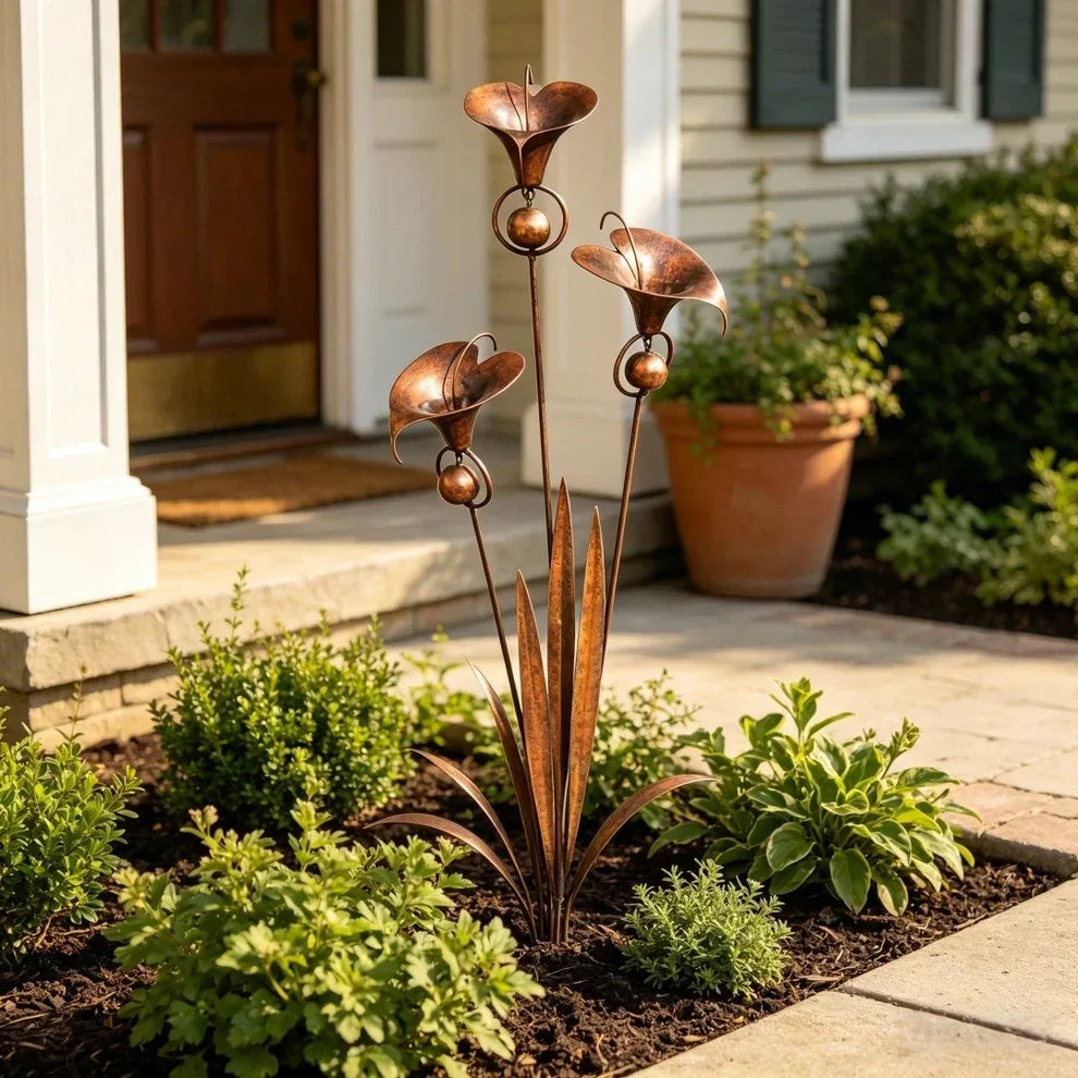 Copper flower stake with three trumpet blooms and bead accents, planted in a mulched bed beside a front porch and terracotta pot. 1