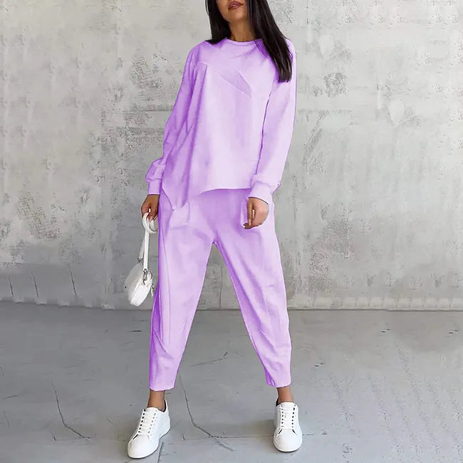 Woman wearing the light purple set, arms relaxed at her sides, holding a white bag in front of a concrete wall.
