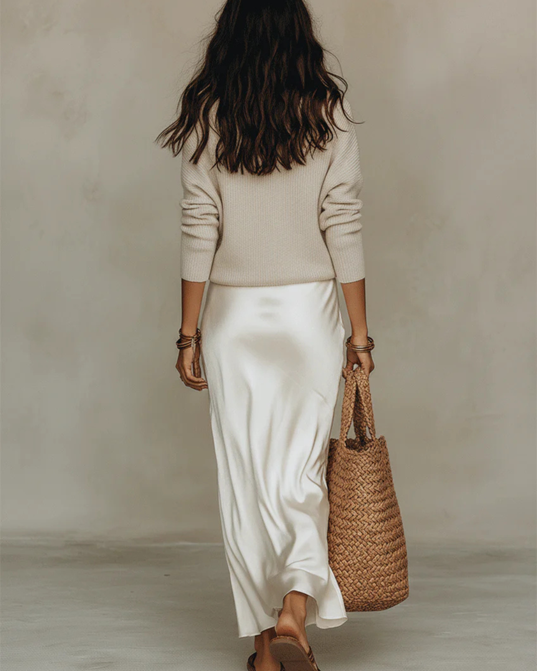 Back view of the model walking away in the cream satin midi skirt and beige knit sweater, carrying a woven straw tote. 9