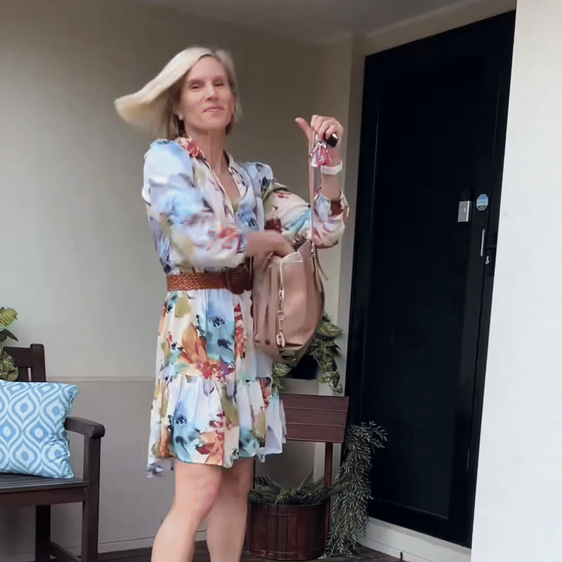 Blonde woman in a floral dress and brown belt tossing her smooth hair as she heads out the front door with her bag.