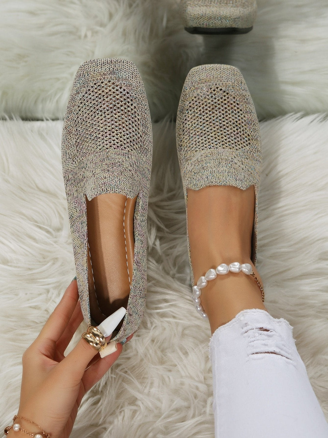 Top-down view of speckled beige knit slip-on flats with perforated mesh toes, one worn on a foot with a pearl anklet, one held by hand. 21