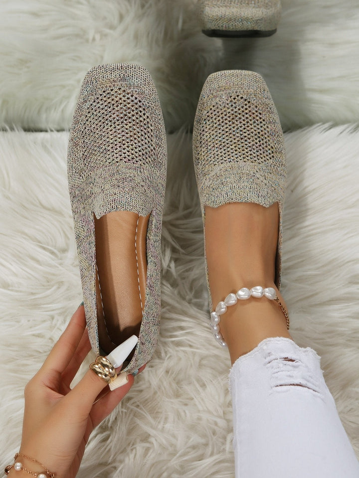 Top-down view of speckled beige knit slip-on flats with perforated mesh toes, one worn on a foot with a pearl anklet, one held by hand. 21
