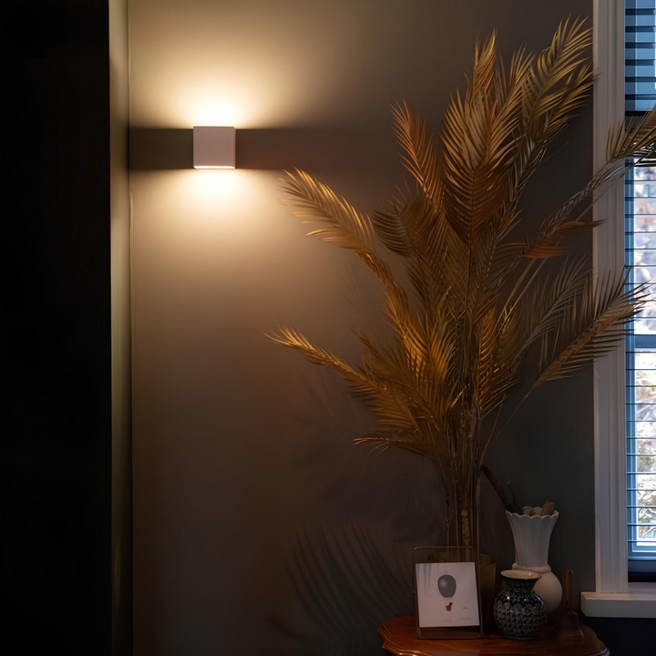 White cube wall lamp glowing on a dark wall in a living room, next to golden palm leaves and a small vase. 4