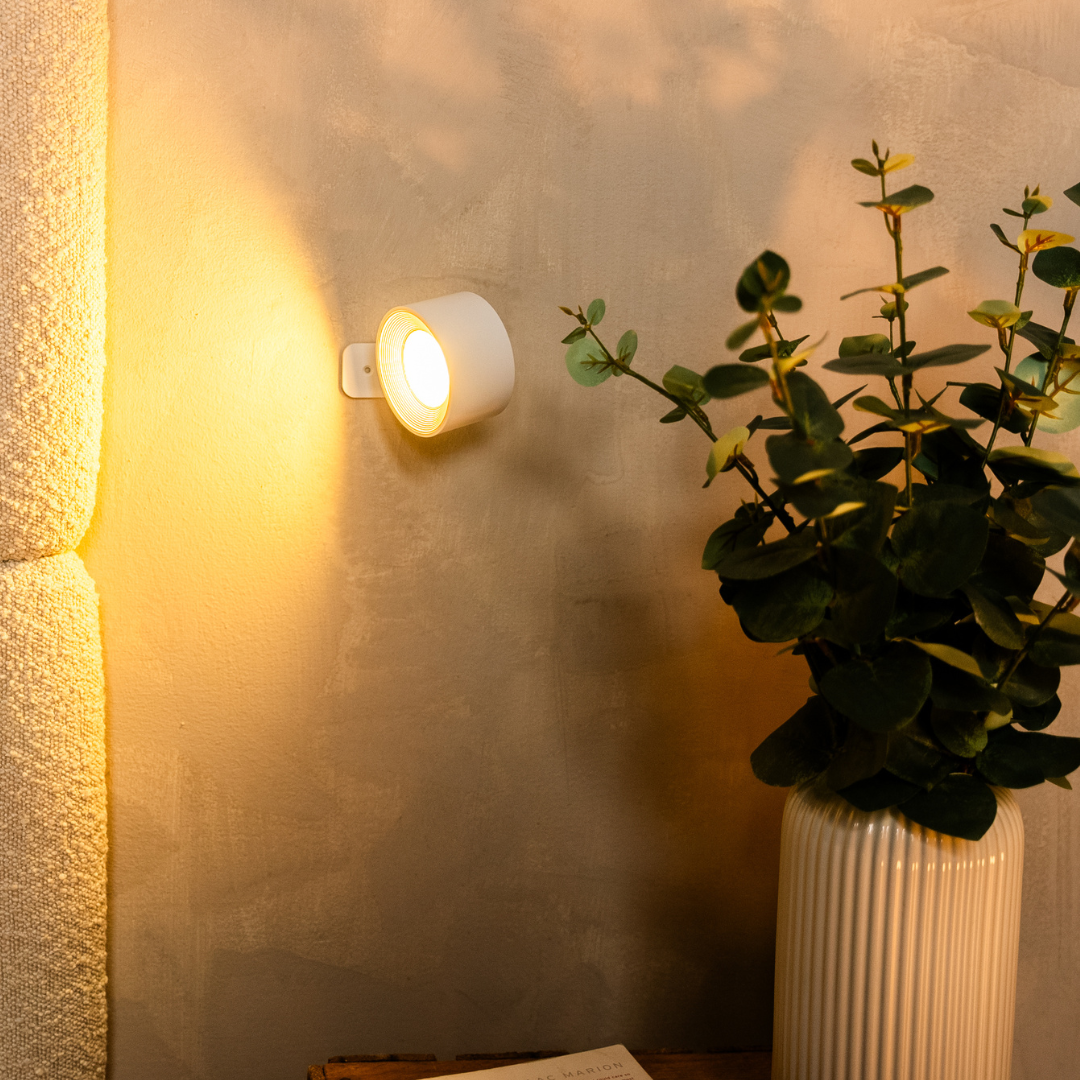 Black wireless LED wall light, another view, glowing beside a boucle headboard and vase.