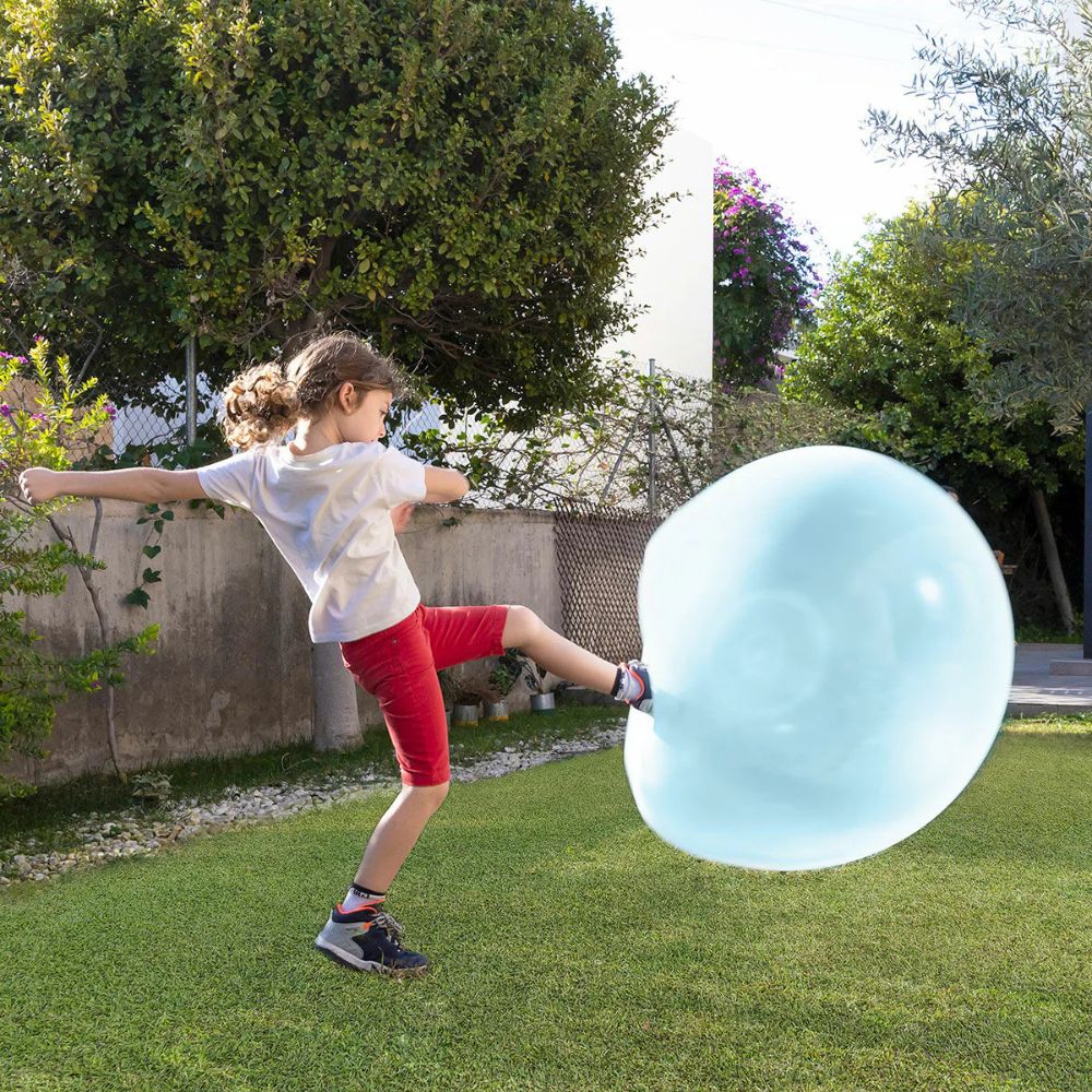Child in white shirt and red shorts kicks a large translucent light blue inflatable ball across a sunny backyard lawn. 4