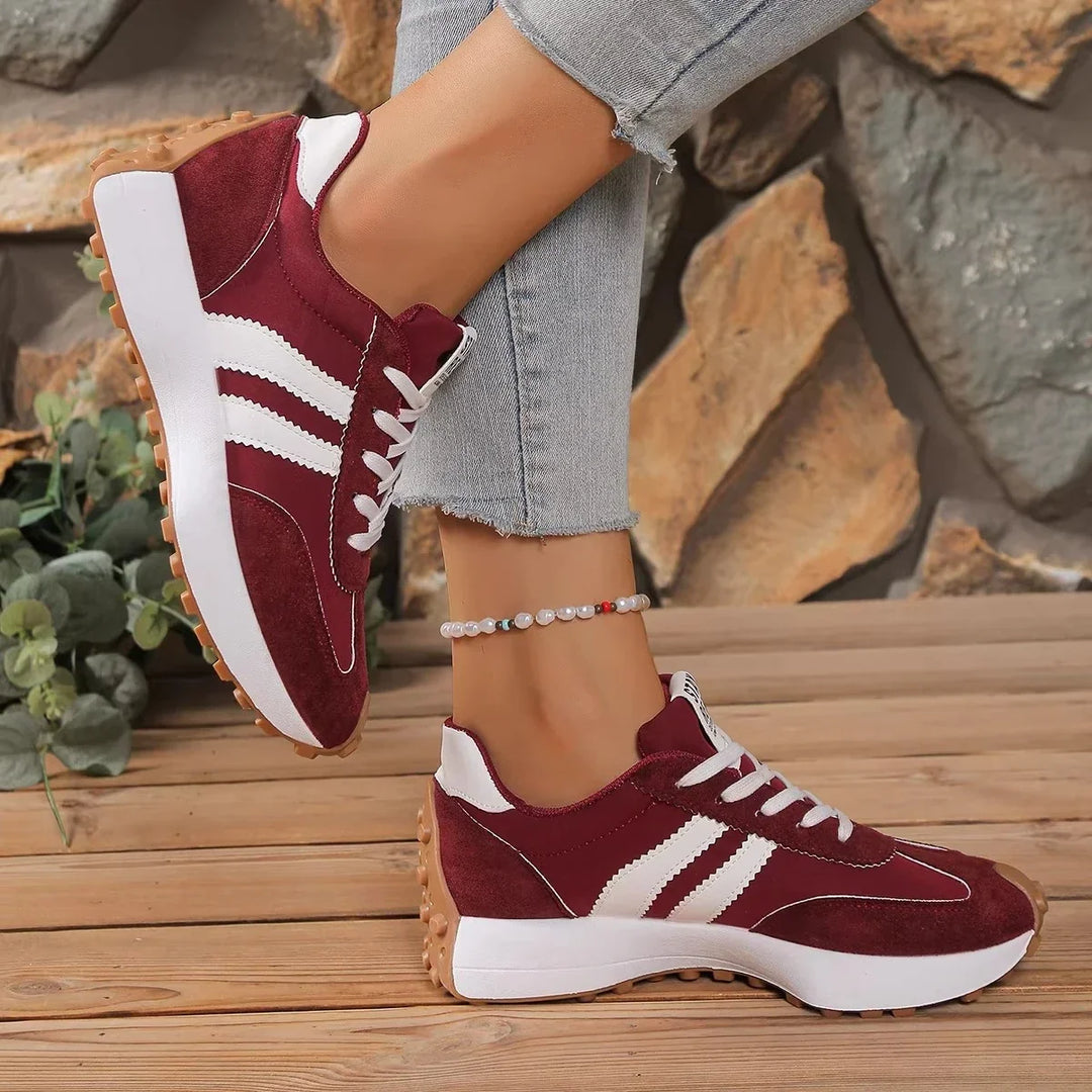 Burgundy trainers with white stripes and a gum-colored sole, worn with one foot raised against a rock wall 5