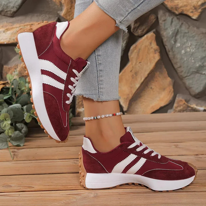 Burgundy trainers with white stripes and a gum-colored sole, worn with one foot raised against a rock wall 5