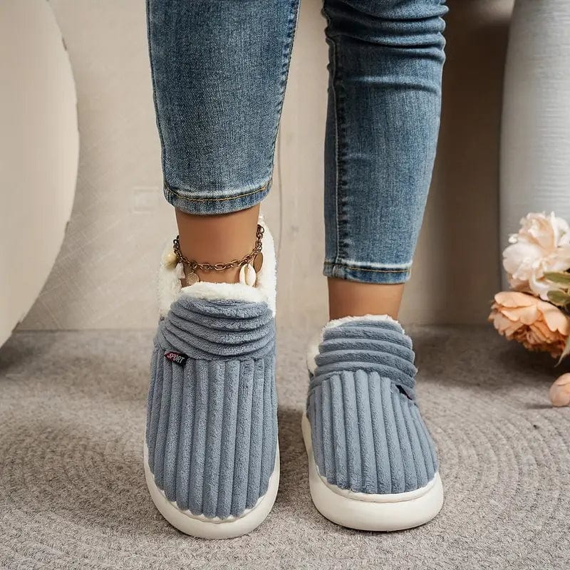 Front view of feet wearing gray-blue ribbed slippers with fleece collars, jeans and a shell anklet, standing on a gray rug.