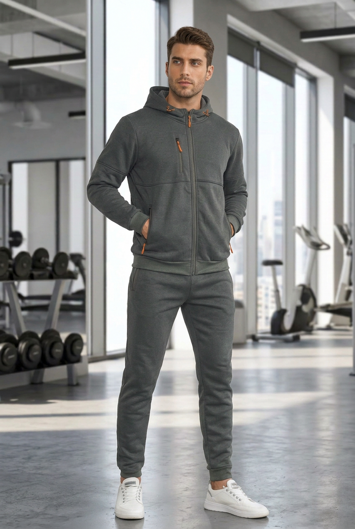 Model in a gray zip-up hoodie and jogger pants with orange zipper pulls, standing in a gym 1