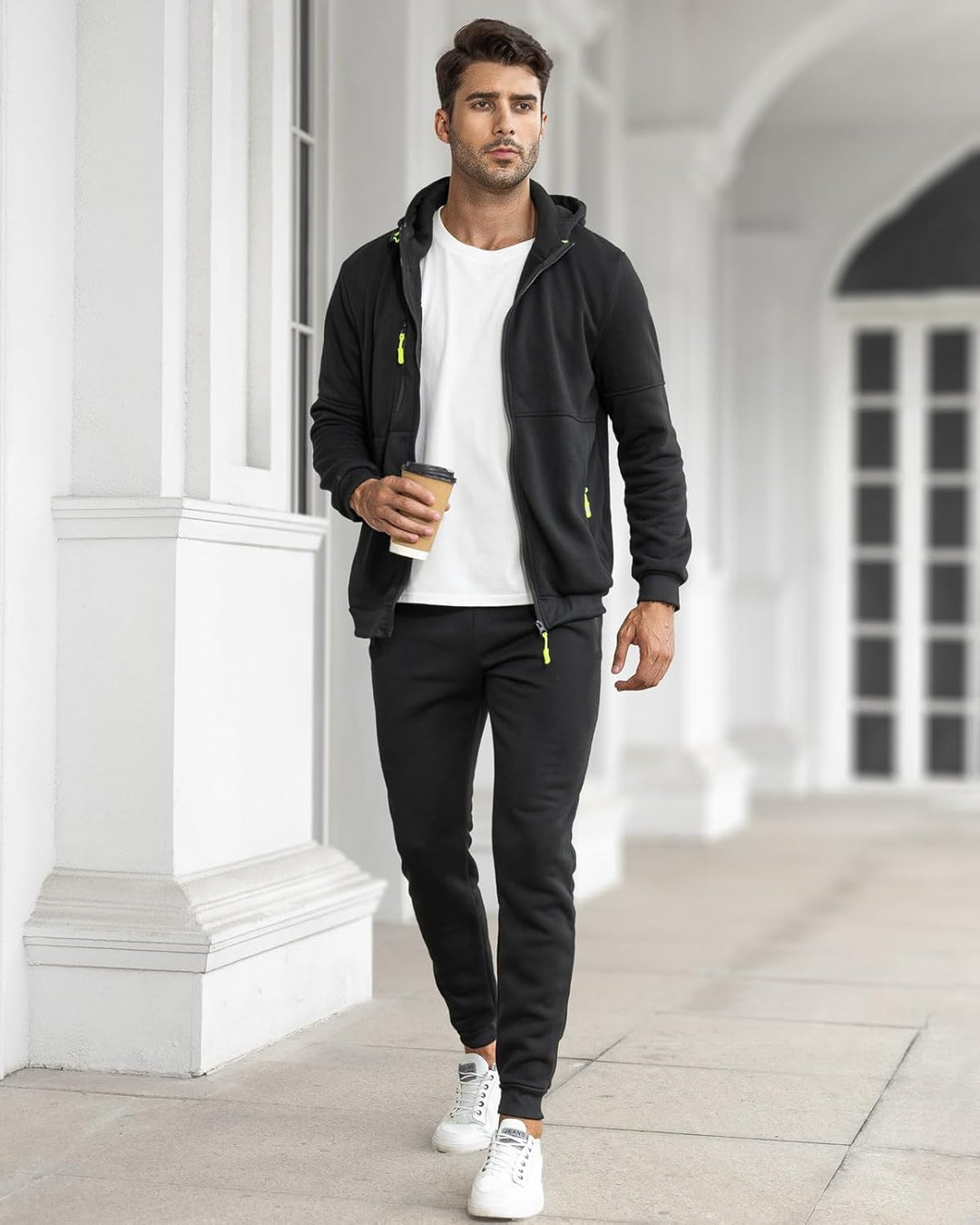 Model walking outdoors in the black tracksuit with the jacket open over a white t-shirt, holding a coffee 25