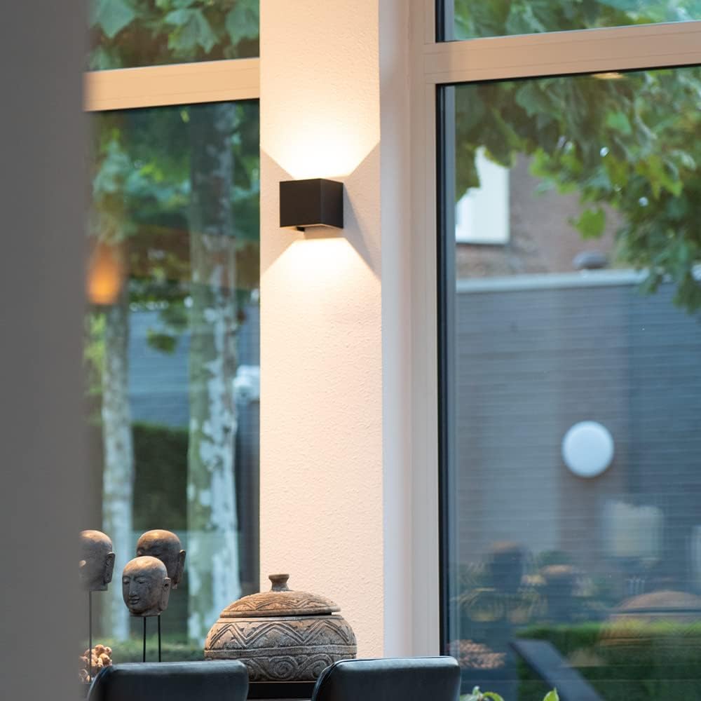 Black cube wall lamp on a white pillar between large windows, glowing above decorative stone heads and a carved lidded bowl. 8