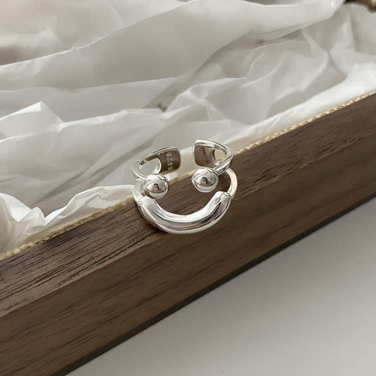 Ring displayed upright in a wooden tray with crumpled white tissue paper, its ball eyes and curved smile facing the camera. 6