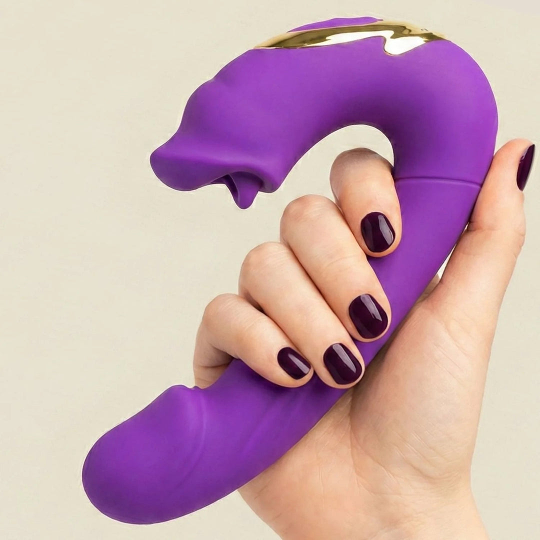 Hand with dark purple nails holding the purple silicone massager, showing its curved shape, gold accent and tongue-like tip against a plain background. 4