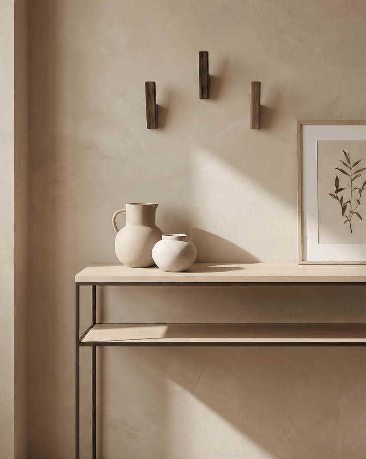 Three dark wood hooks mounted in a triangle above a slim console table with cream ceramic vases and a framed botanical print. 3