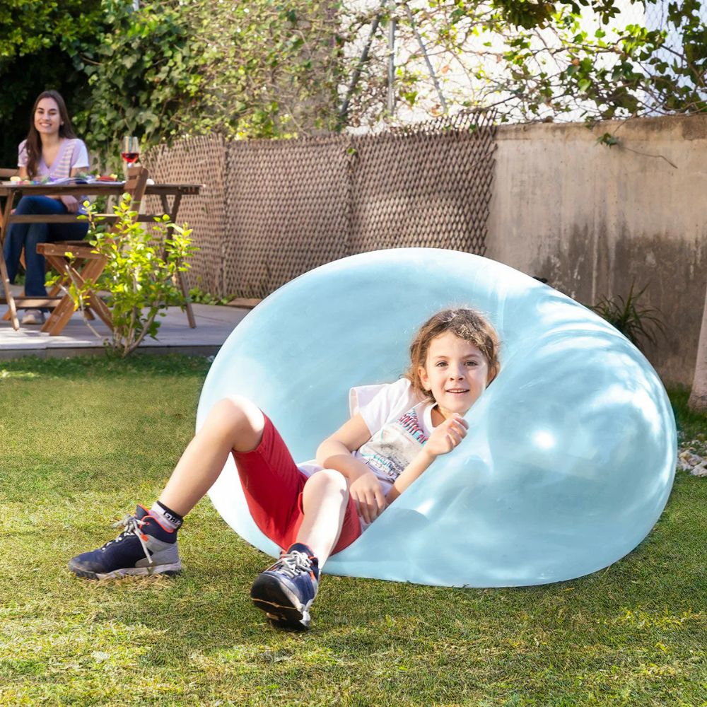 Smiling child in red shorts reclines against a large translucent light blue inflated ball on a backyard lawn while a woman watches from a patio table. 5