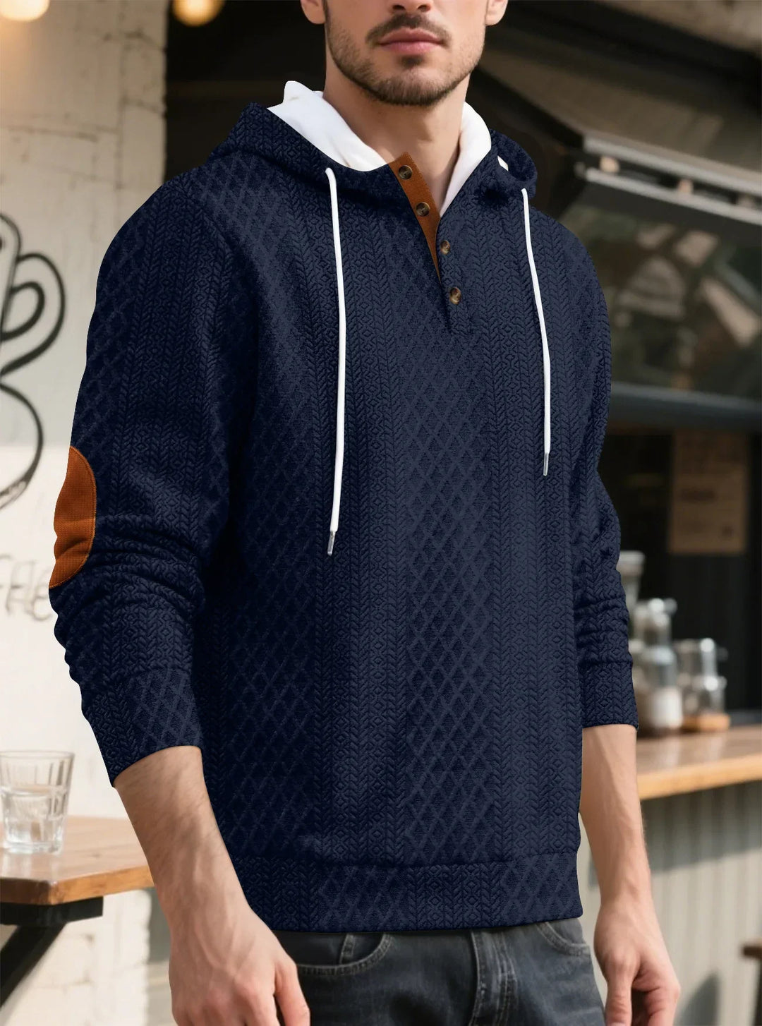 Man wearing a navy diamond-textured hoodie with white drawstrings and brown elbow patch, standing near a cafe counter 6