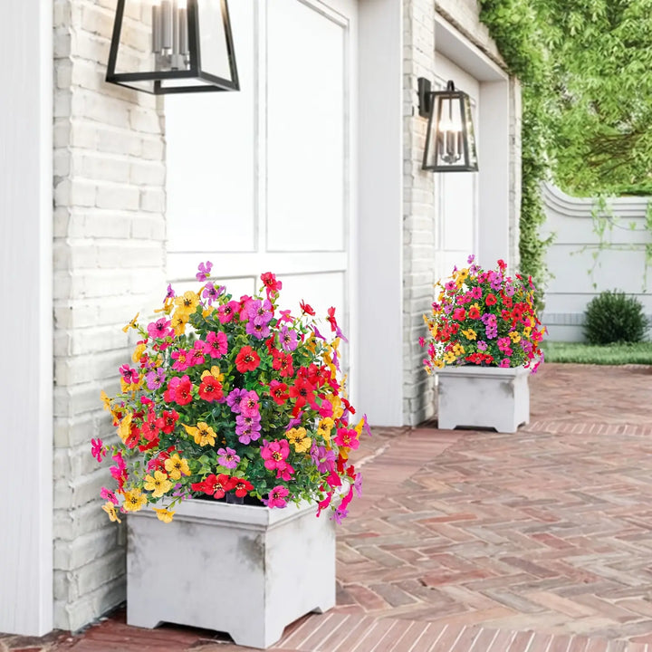 Multicolor artificial flower bushes in white square planters on a brick patio.