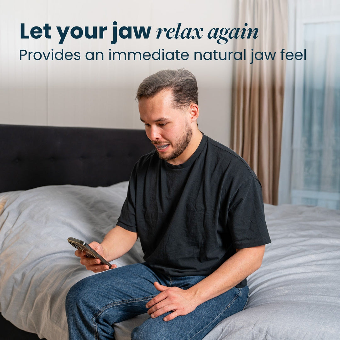Man in a black T-shirt sits on the edge of a bed looking at his phone with the mouthpiece in his mouth. 26
