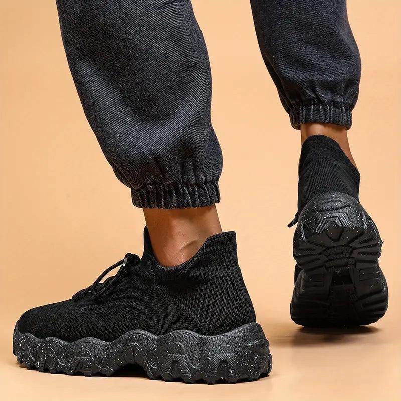 Another view of black knit sneakers from the rear on a model in gray joggers, with one sole raised to show the tread.