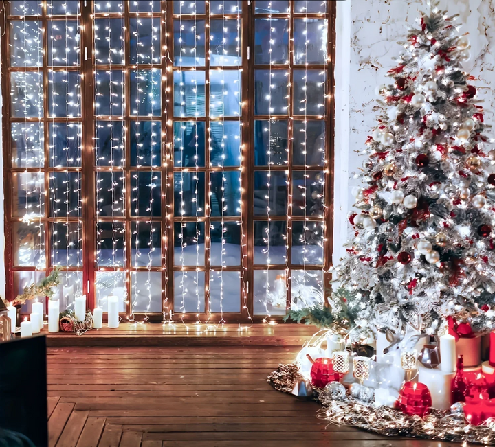Cool white curtain lights covering a large paned window, next to a frosted Christmas tree with red ornaments and candles. 5