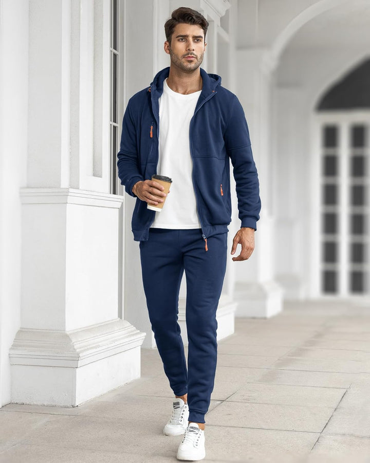 Model walking outdoors in the navy tracksuit with the jacket open over a white T-shirt, holding a coffee cup 7