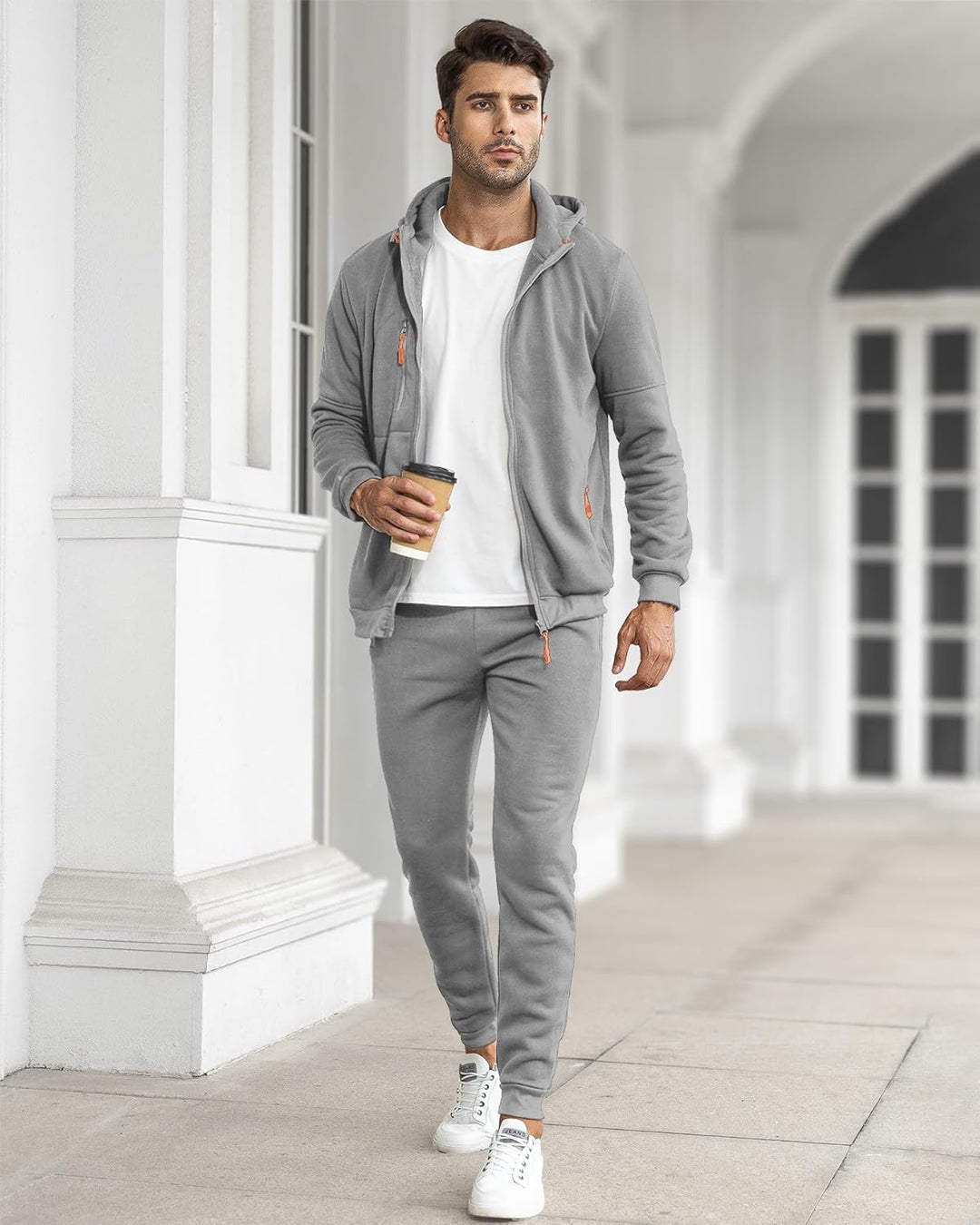 Model walking outdoors in the gray tracksuit with the jacket open over a white t-shirt, holding a coffee 31