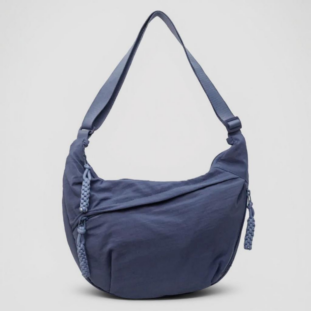 Front view of a blue crescent-shaped fabric shoulder bag with an adjustable strap, diagonal zipper, and braided zipper pulls on a plain background. 1
