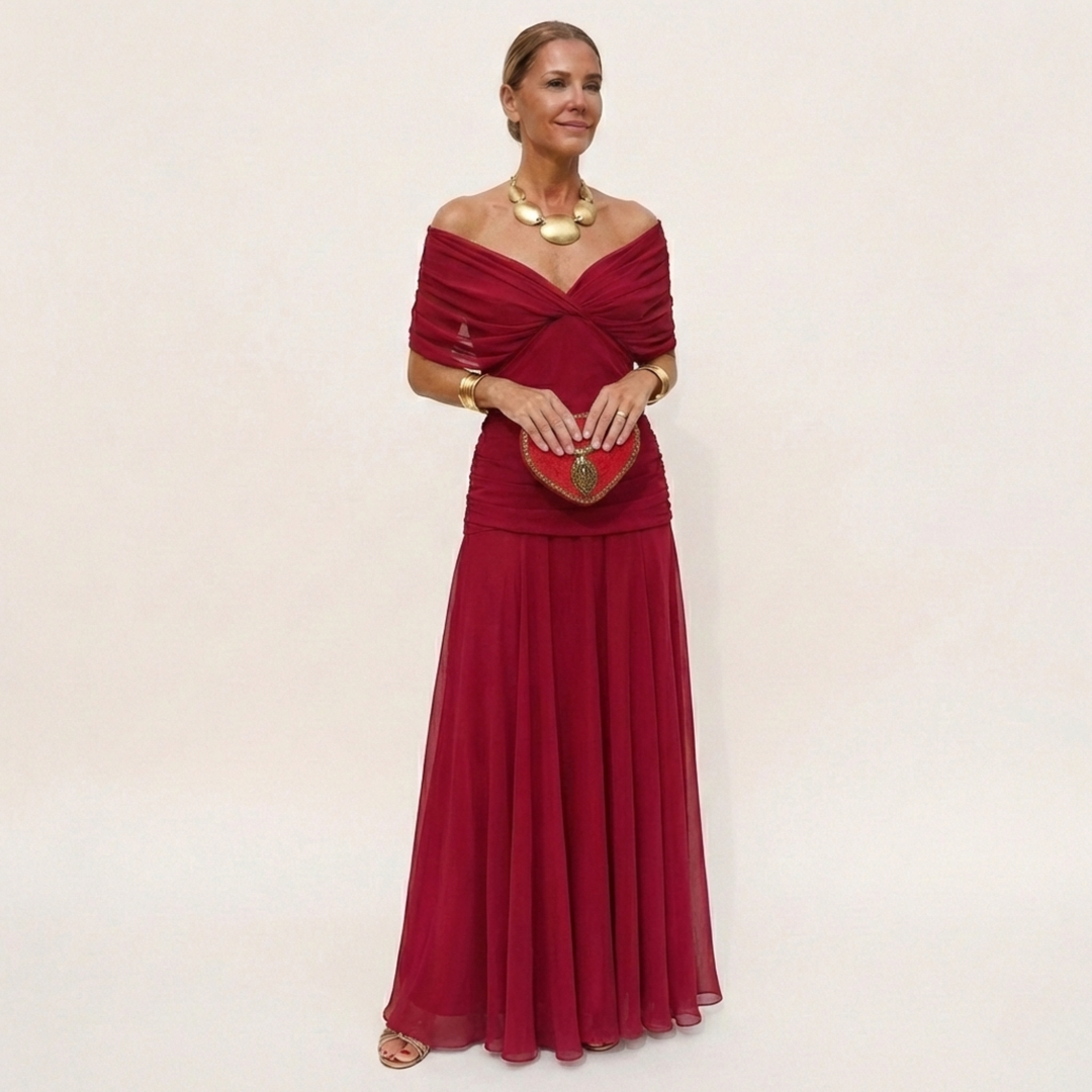 Model in a red off-the-shoulder maxi dress with a twisted neckline and ruched waist, holding a red heart-shaped clutch. 5