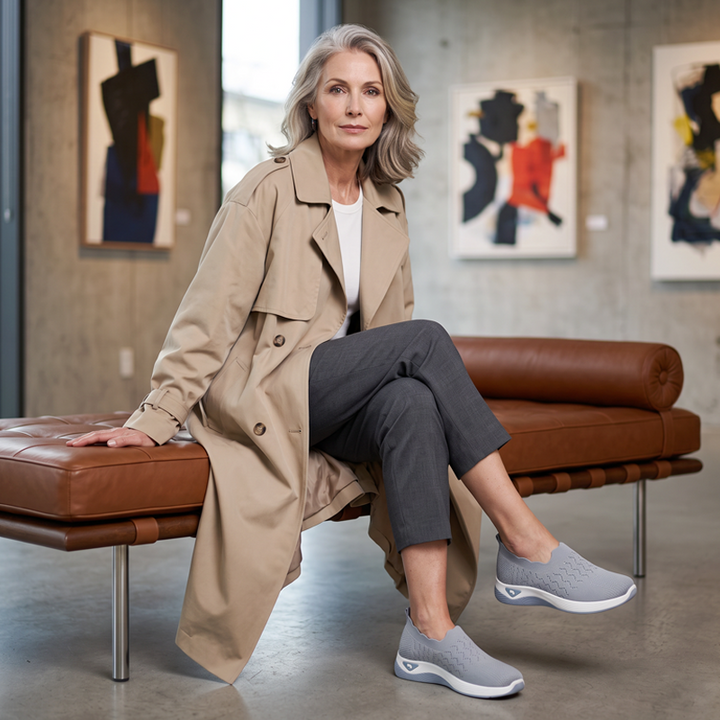 Older woman in a beige trench coat sits on a leather bench wearing gray slip-on sneakers.