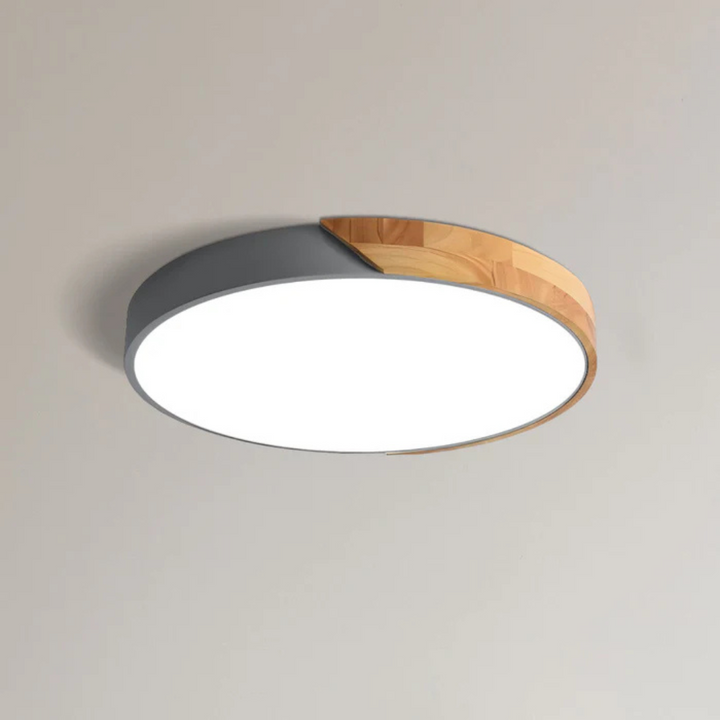 Gray round ceiling light with a natural wood section on the rim, mounted on a light ceiling and glowing white.
