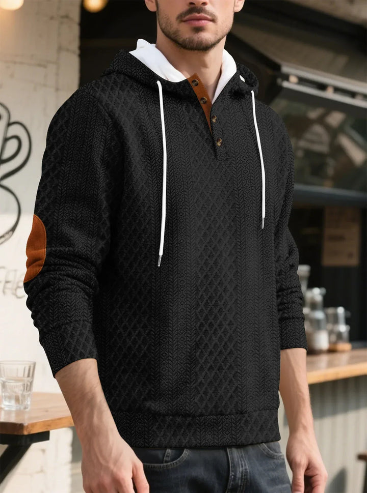 Man wearing a black diamond-textured hoodie with white drawstrings and brown elbow patch, standing near a cafe counter 7